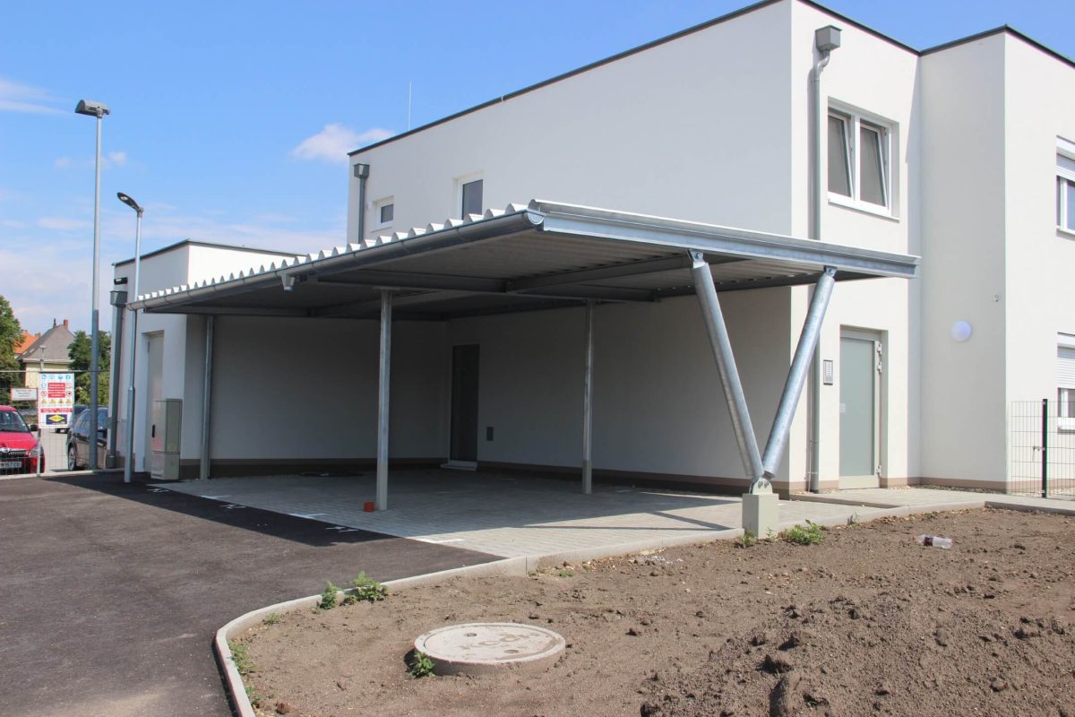 Moderner Carport aus verzinktem Stahl mit markanter V-Stütze, integriert in ein schlichtes, weißes Wohngebäude – idealer Schutz für zwei Fahrzeuge.