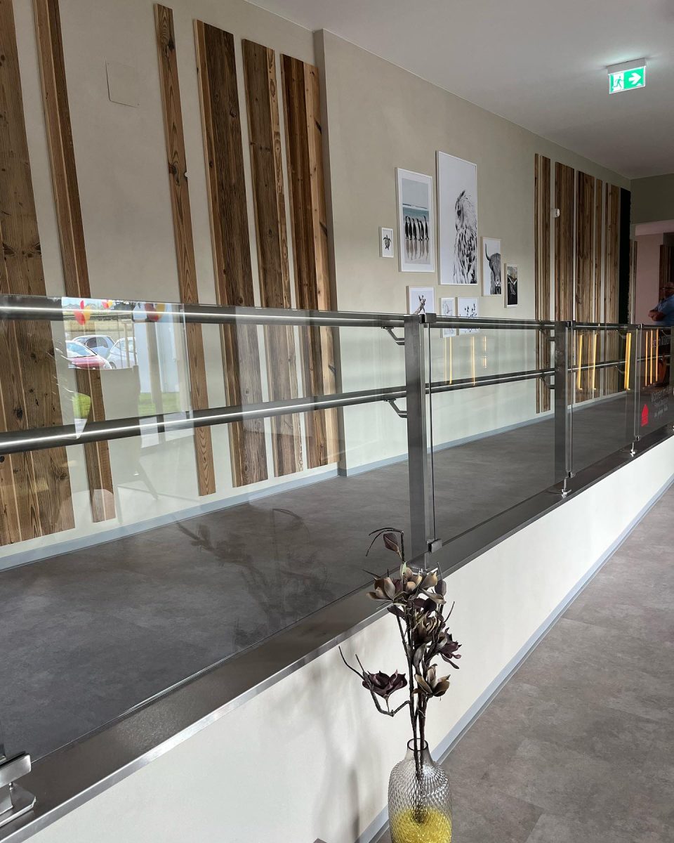Modernes Geländer mit Glasfüllung und Edelstahlpfosten im Innenbereich eines Gebäudes mit dekorativer Wandgestaltung und Notausgangsschild
