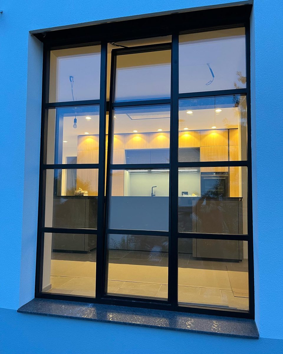 Großes Sprossenfenster mit schwarzem Stahlrahmen in moderner Fassade, Blick in eine beleuchtete Küche