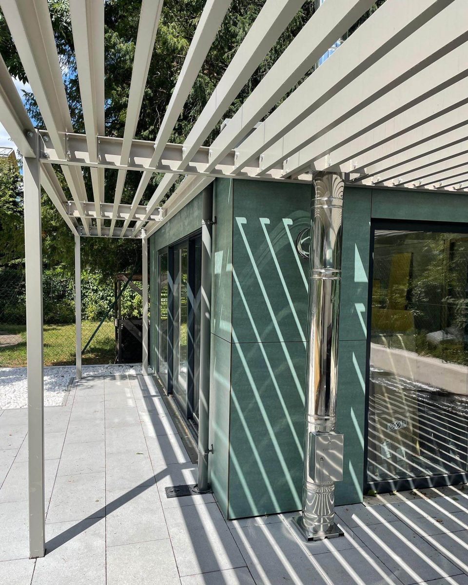 Moderne Pergola aus Metall mit Lamellenüberdachung, die Schattenmuster auf Terrasse und Fassade wirft