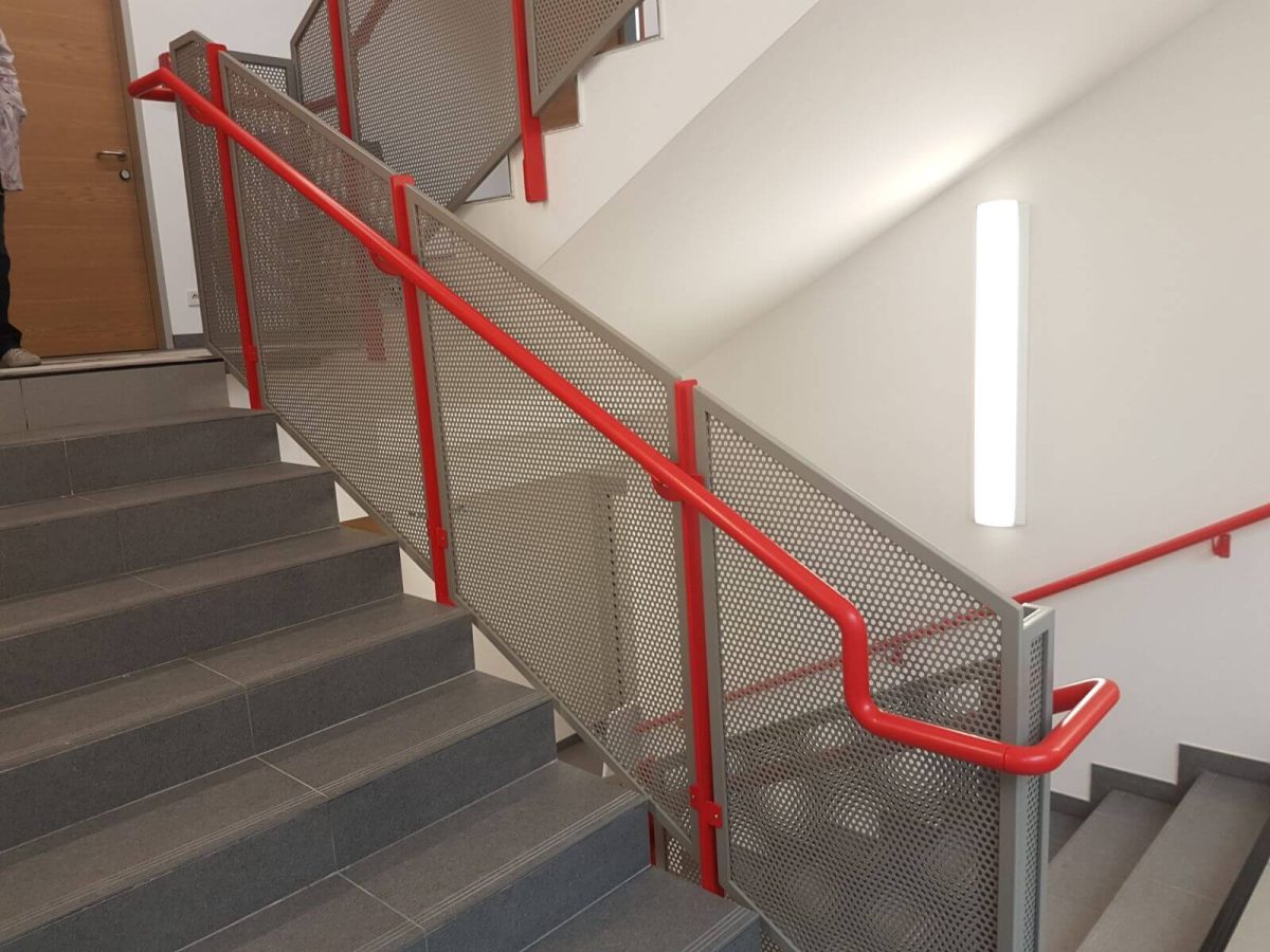 Treppengeländer mit robustem Rahmen aus pulverbeschichtetem Stahl, gefüllt mit Lochblechplatten und auffälligem, rotem Handlauf – ideal für funktionale und öffentliche Bereiche