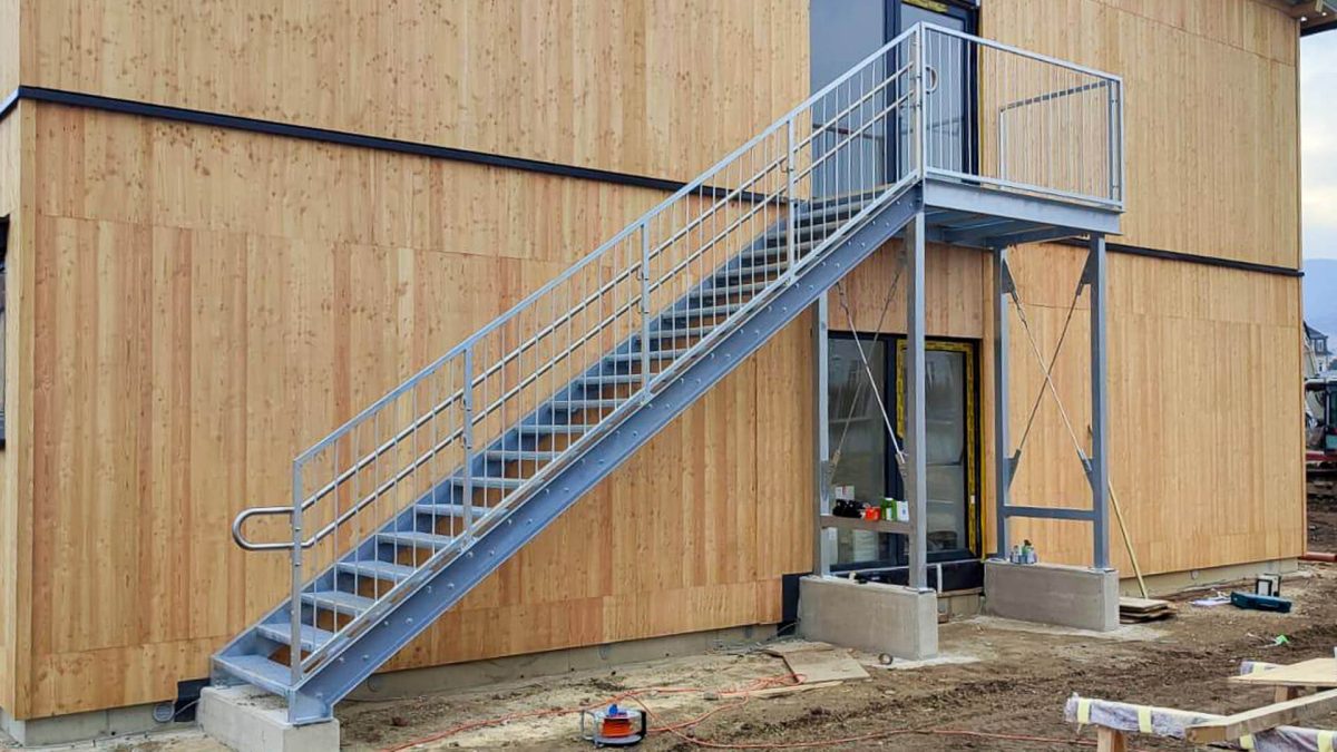 Außentreppe aus verzinktem Stahl mit geradem Aufgang, beidseitigem Geländer und Zwischenpodest, montiert an einem Gebäude mit vertikaler Holzfassade während der Bauphase