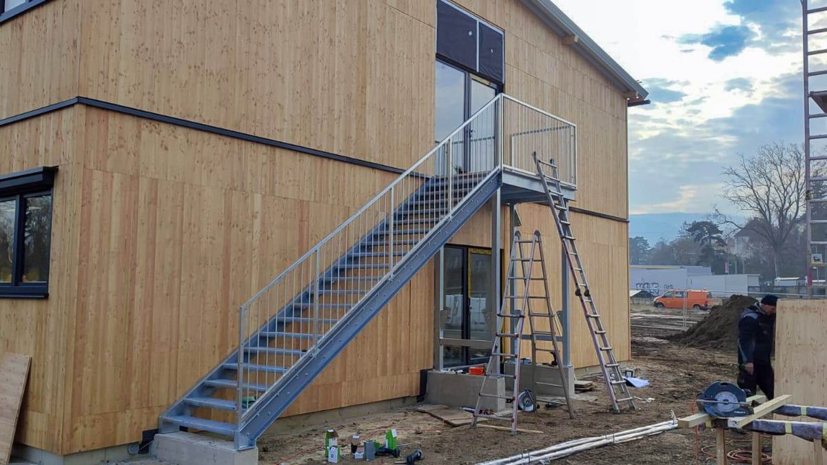Geradläufige Außentreppe aus verzinktem Stahl mit Gitterroststufen und senkrechtem Geländer, montiert an einem modernen Gebäude mit vertikaler Holzfassade und Baustellensituation