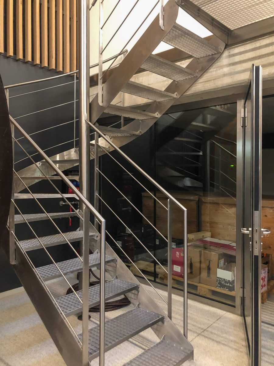 Moderne Spindeltreppe aus Edelstahl mit rutschfesten Gitterroststufen und horizontalen Seilfüllungen, eingebaut in einen Innenraum mit Glasfront und Sichtbetonelementen