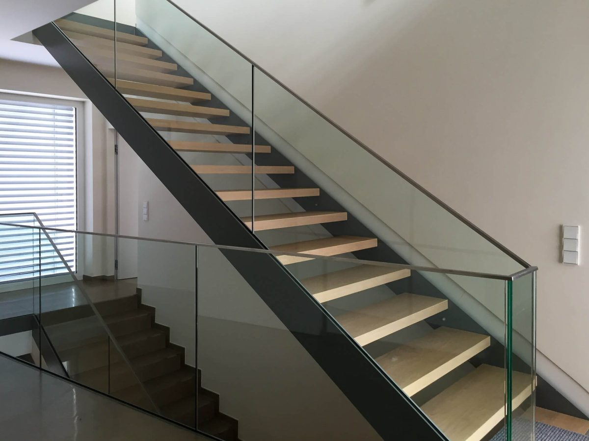 Elegante, freitragende Innentreppe mit hellen Holzstufen und seitlich montiertem Glasgeländer, kombiniert mit schwarzer Stahlwange in einem modernen Wohnambiente