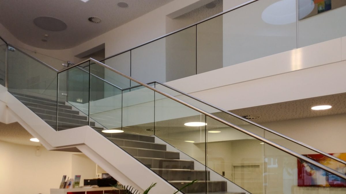 Innenraumtreppe mit glattem Stahlträger, belegt mit Stein- oder Feinsteinzeugstufen, ausgestattet mit einem durchgängigen Ganzglasgeländer und Edelstahl-Handlauf – elegantes Design für öffentliche oder gewerbliche Gebäude