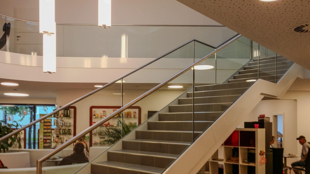 Innenarchitektur mit moderner Treppe in einem öffentlichen Gebäude, ausgestattet mit Glasgeländer und Edelstahlhandlauf für ein offenes, helles Ambiente.