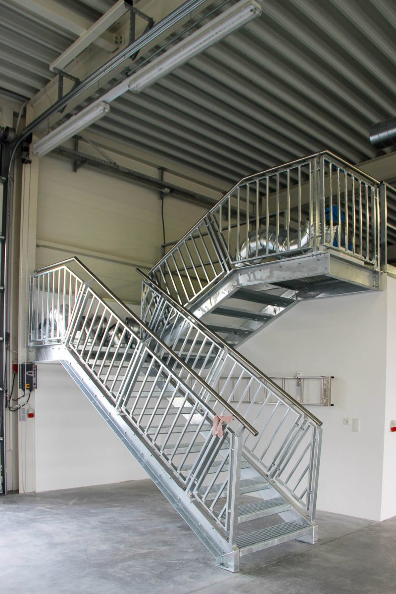 Zweistufige Industrietreppe aus feuerverzinktem Stahl mit Gitterroststufen und stabilem Geländer, montiert in einer Werkhalle mit Sichtdachkonstruktion