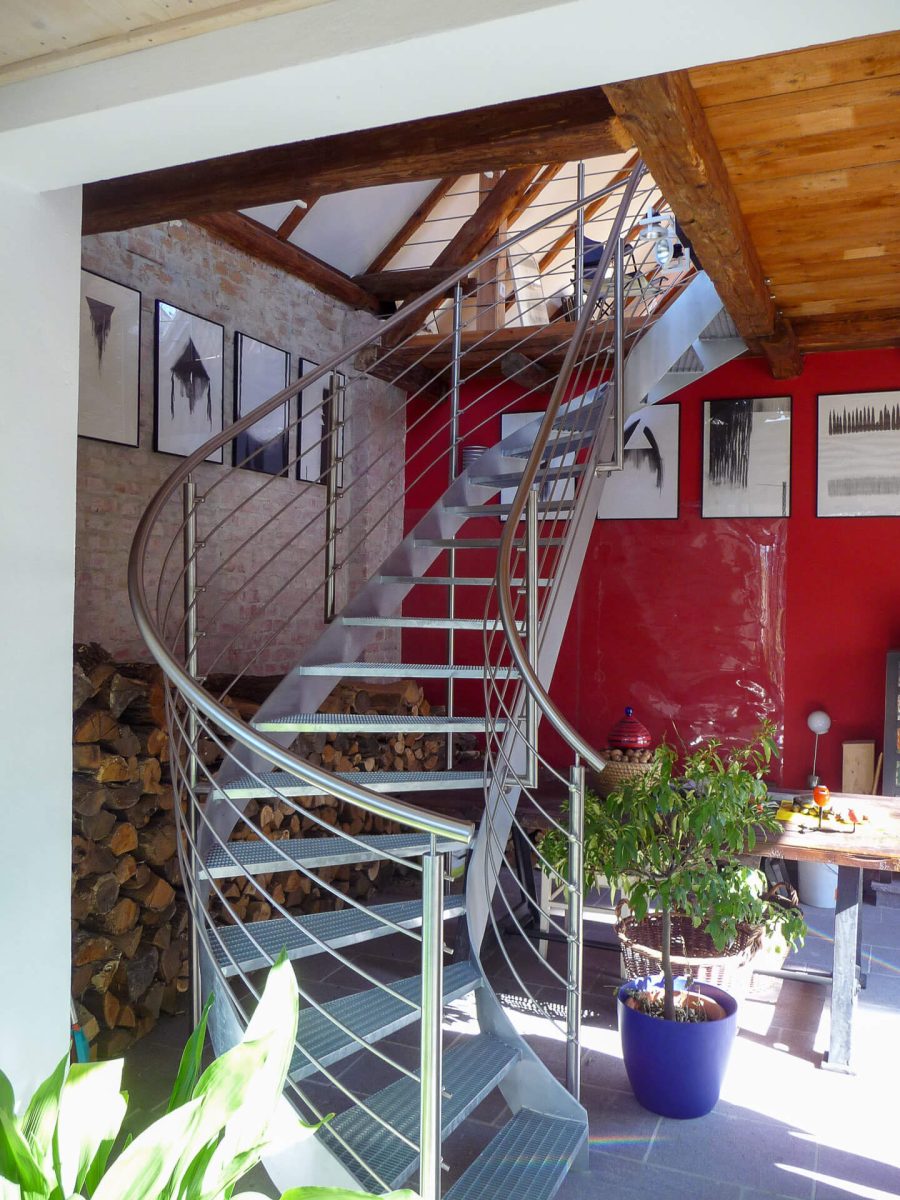 Moderne gewendelte Stahltreppe mit rutschfesten Gitterroststufen und elegantem Edelstahlgeländer, eingebaut in einen Wohnraum mit sichtbaren Holzbalken und Backsteinwand