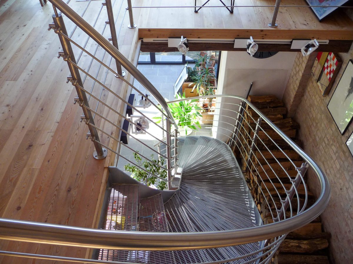 Moderne Wendeltreppe mit Gitterroststufen und geschwungenem Edelstahlgeländer, eingebaut in einen offenen Wohnbereich mit Holzfußboden und Galerieebene