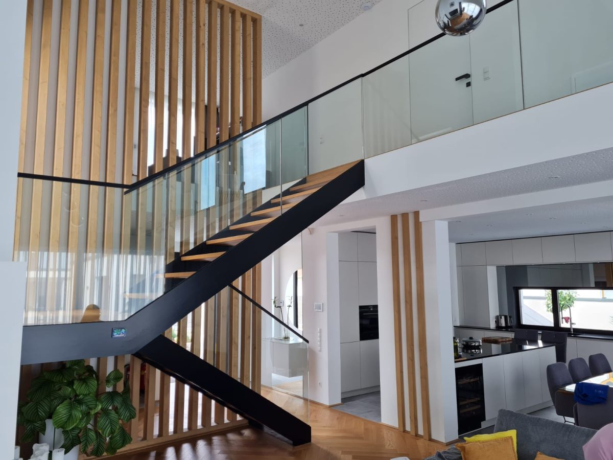 Moderne Innentreppe mit schwarzem Stahlträger, Holzstufen und Glasgeländer in einem offenen Wohnraum mit Sicht auf Küche und Galerie.