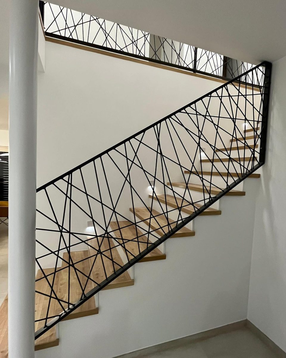 Schräg verlaufendes Treppengeländer aus schwarzem Stahl mit unregelmäßig gekreuzten Streben, kombiniert mit Holzstufen und weißer Wand, in einem modernen Innenraum