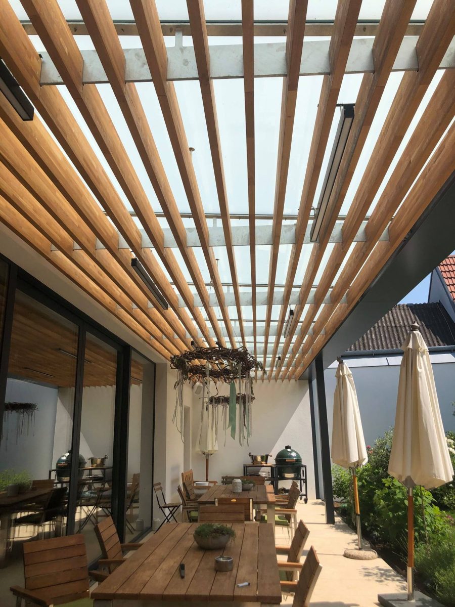 Moderne Terrassenüberdachung mit Glasdach und Holzlamellen, darunter gemütliche Sitzbereiche mit Holztischen und dekorativen Elementen.