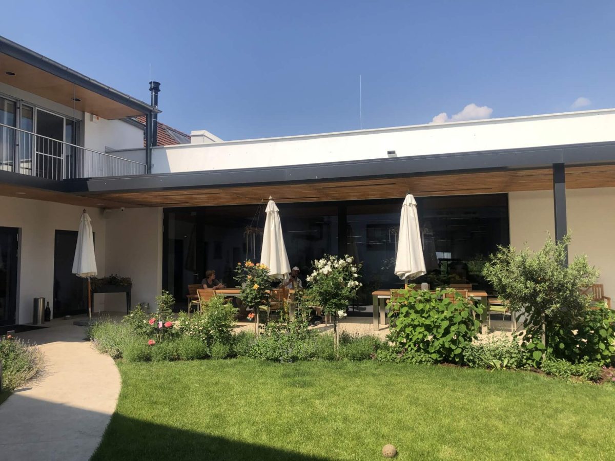 Grüner Innenhof mit gepflegtem Rasen, Blumenbeeten und überdachter Terrasse mit Sitzgruppen und Sonnenschirmen – ideal für entspannte Zusammenkünfte.