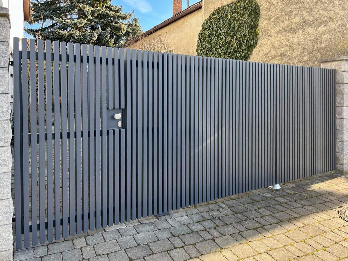 Langer Sichtschutzzaun aus dunkelgrauen, vertikalen Aluminiumlamellen mit integrierter Tür und Türgriff, montiert zwischen massiven Steinsäulen auf einem gepflasterten Vorplatz