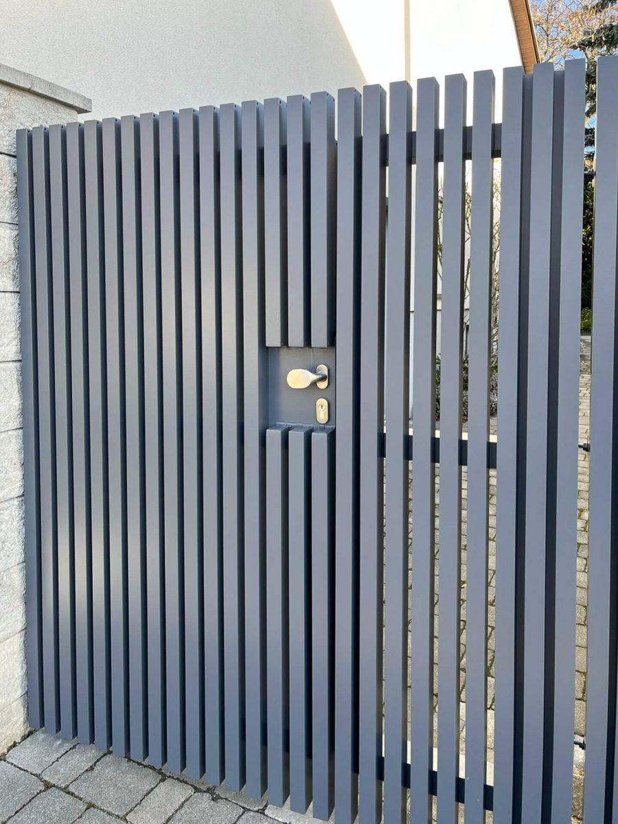 Nahaufnahme eines modernen Aluminiumzauns mit vertikalen Lamellen und integrierter Tür, inklusive Türgriff und Zylinderschloss, montiert zwischen Steinsäulen