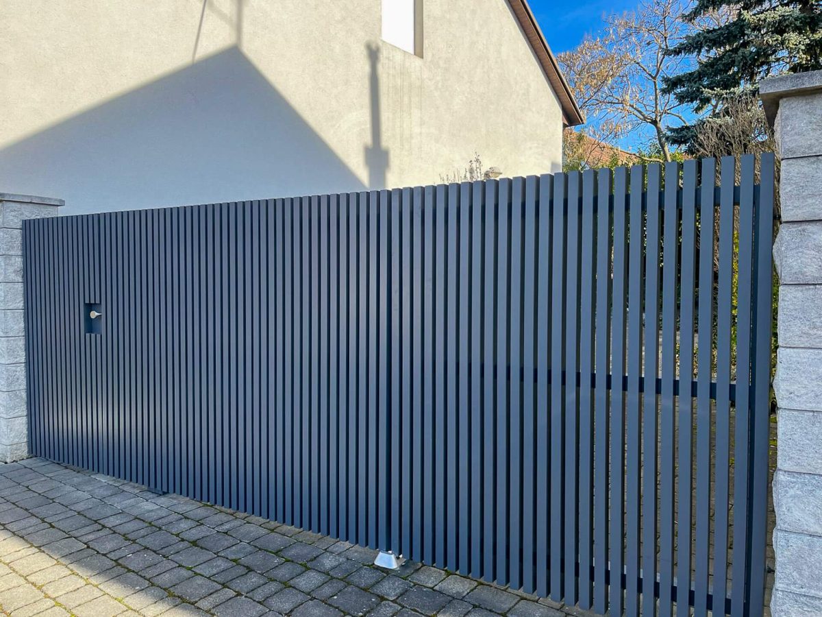 Moderner Sichtschutzzaun aus dunkelgrauen, vertikal angeordneten Aluminiumlamellen mit integrierter Tür, eingefasst zwischen Steinsäulen auf gepflastertem Boden