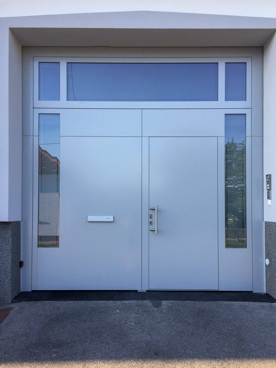 Schlichtes graues Einfahrtstor mit integrierter Tür, klarer Linienführung und seitlichen sowie oberen Glaselementen, eingebettet in eine helle Putzfassade mit Granitsockel