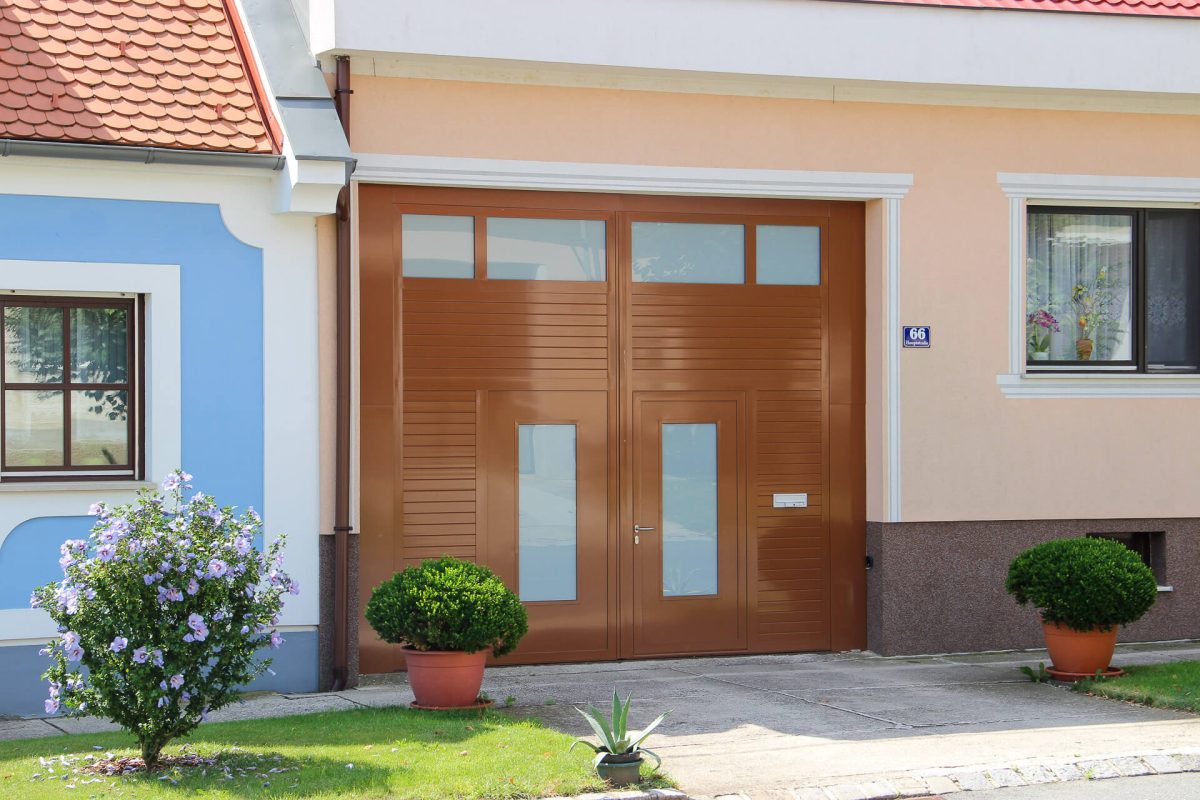 Modernes Einfahrtstor in brauner Holzoptik mit integrierter Tür und Milchglas-Elementen, harmonisch eingefasst zwischen zwei Wohnhäusern mit gepflegtem Vorgarten