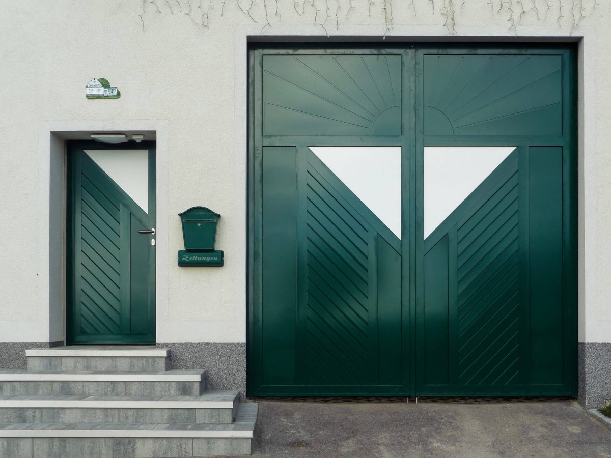 Grünes Einfahrtstor mit geometrischem Muster und passender Haustür, beide mit weißen Glaseinsätzen und diagonal verlaufenden Lamellen, vor hellem Putz und grauen Stufen