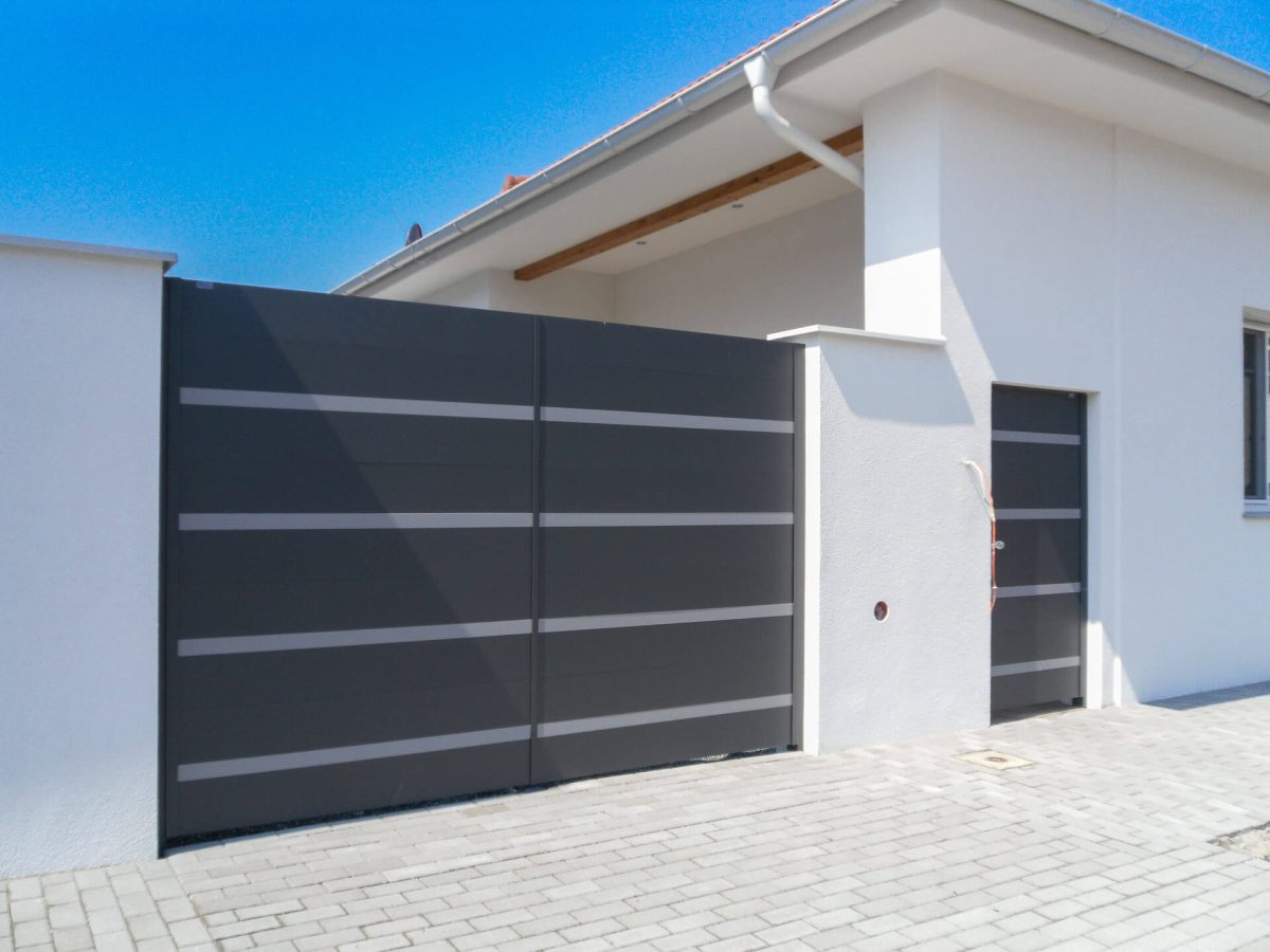 Zeitgemäßes Einfahrtstor in Dunkelgrau mit dezenten horizontalen Aluminiumstreifen, ergänzt durch eine passende Nebentür, montiert an einem weißen Neubau mit Pflasterzufahrt
