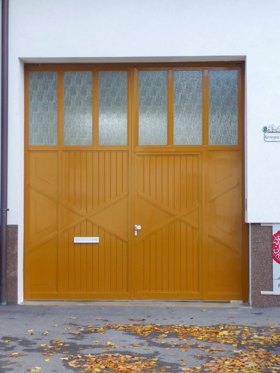 Großes zweiflügeliges Einfahrtstor in warmem Holzton mit integrierter Tür, senkrechten und diagonalen Lamellen sowie einem oberen Segment aus strukturiertem Glas, eingefasst in eine weiße Fassade