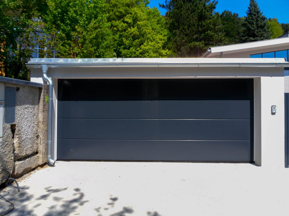 Modernes Garagentor als Sektionaltor in Anthrazitgrau mit glatter Oberfläche und horizontalen Segmenten, eingebaut in eine weiße Garage mit Flachdach