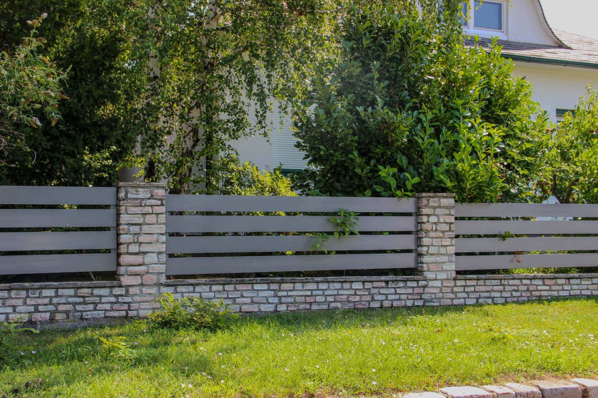 Gartenzaun aus grauen horizontalen Metalllamellen und gemauerten Pfeilern vor dichter Bepflanzung und einem Wohnhaus
