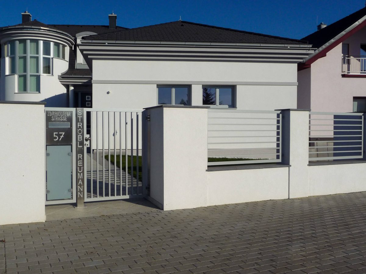 Moderner Gartenzaun aus horizontalen Aluminiumlamellen mit integrierter Tür und Briefkasten, kombiniert mit dunklen Putzpfeilern und -sockel, vor einem Wohnhaus