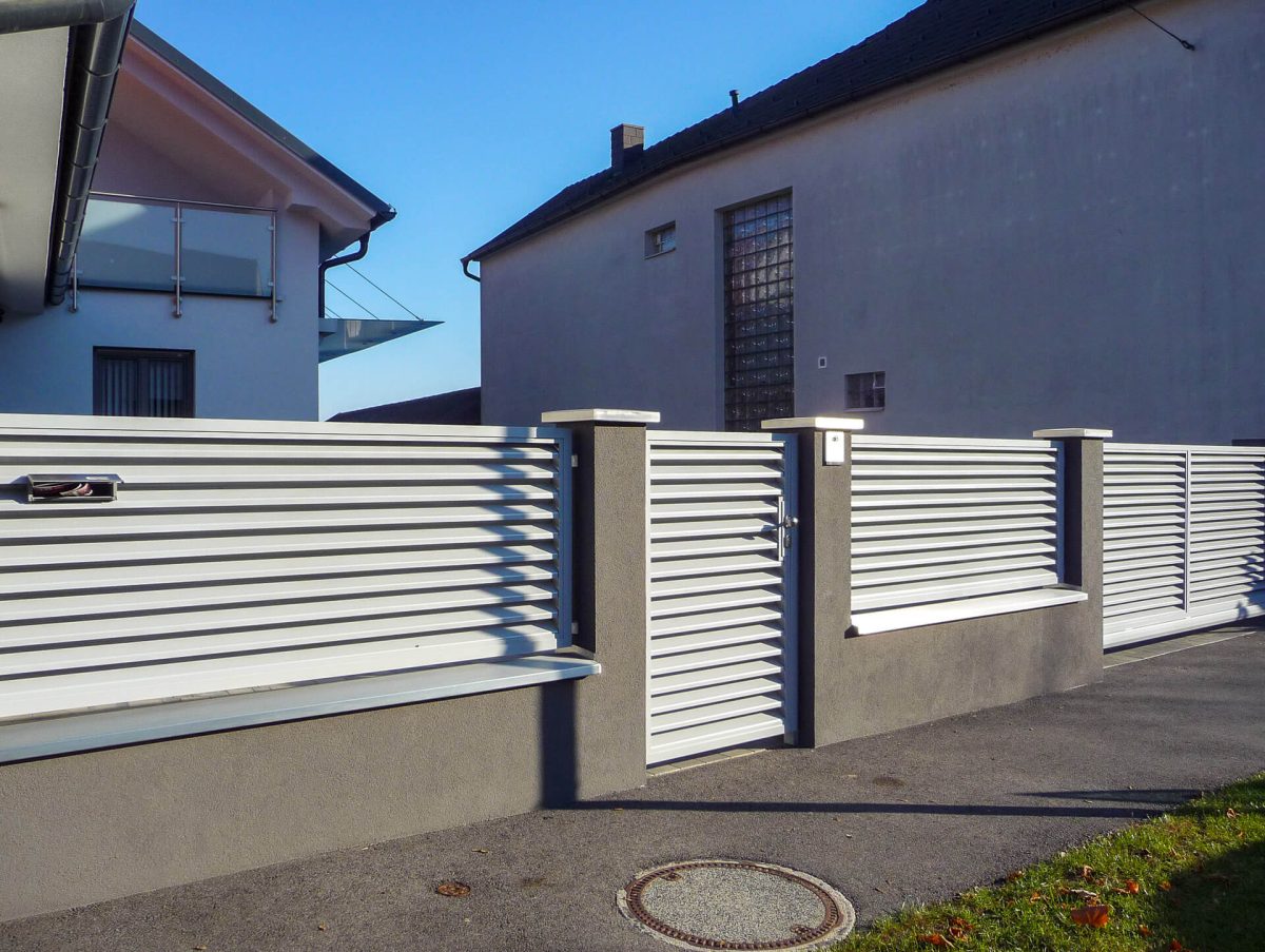 Moderner Gartenzaun aus horizontalen Aluminiumlamellen mit integrierter Tür und Briefkasten, kombiniert mit dunklen Putzpfeilern und -sockel, vor einem Wohnhaus