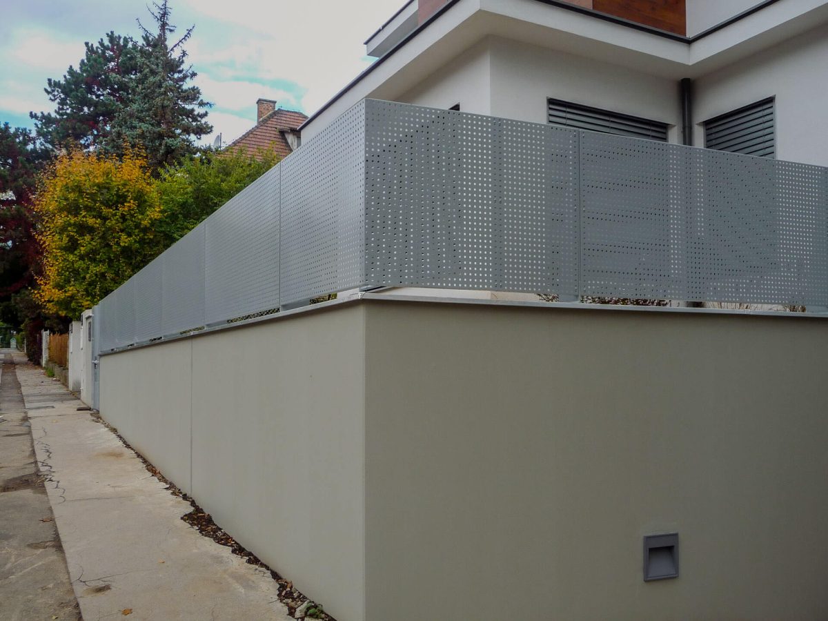 Moderner Sichtschutz aus gelochtem Aluminiumblech auf einem hohen, glatten Betonsockel entlang eines Gehwegs, angrenzend an ein zeitgenössisches Wohnhaus mit Flachdach