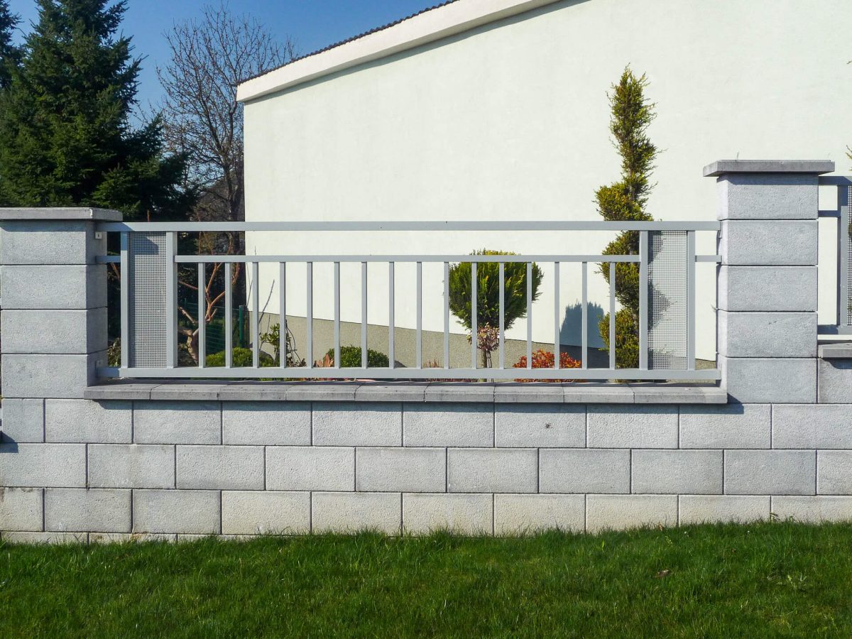 Moderner Gartenzaun aus vertikalen Aluminiumstreben mit seitlichen Lochblecheinsätzen, eingefasst von grauen Betonpfeilern und -sockel vor einer gepflegten Hecke und weißer Hauswand
