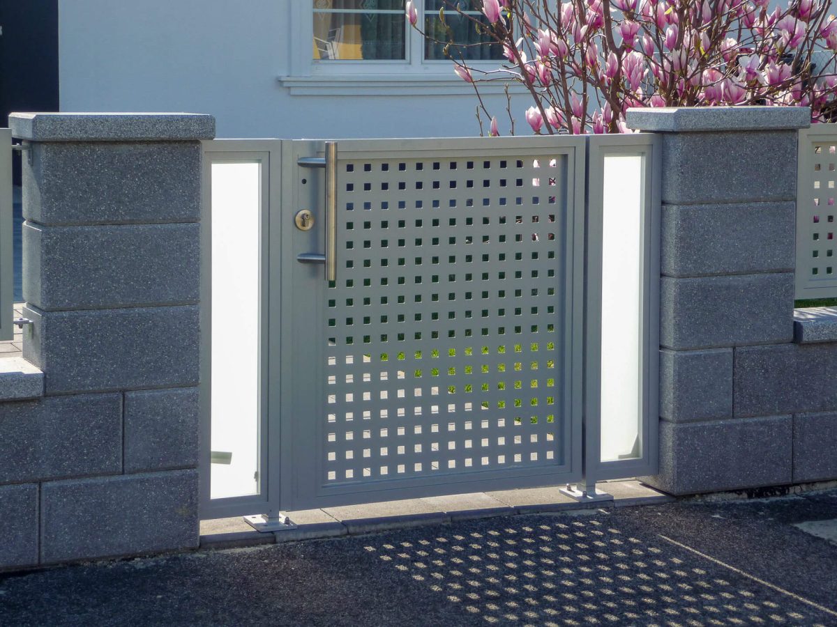 Silbernes Eingangstor mit quadratischer Lochblechfüllung, seitlichen Glasfeldern und massivem Metallgriff, eingefasst von grauen Betonsteinen und blühendem Magnolienbaum im Hintergrund