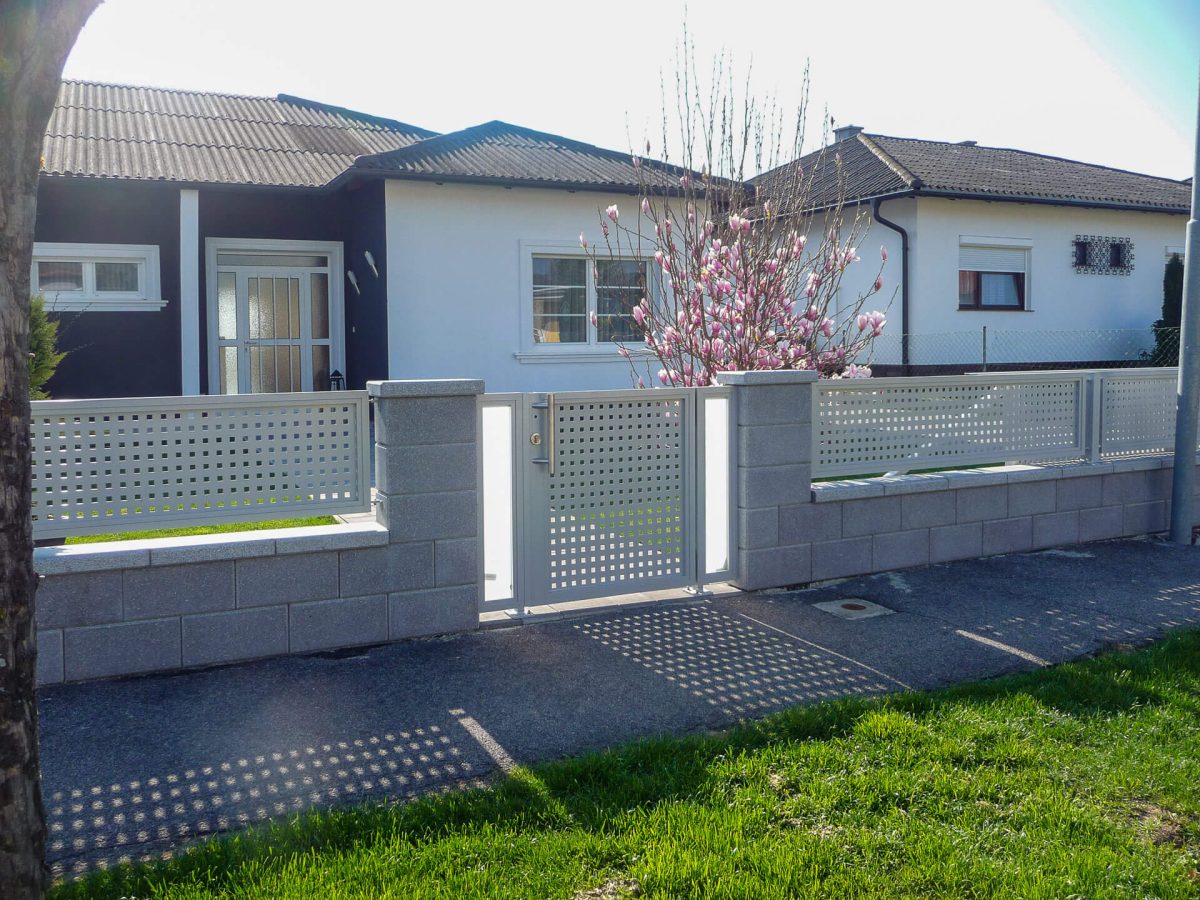 Moderner Gartenzaun mit Tür, bestehend aus Betonsteinen und silberfarbenem Lochblech, vor einem Einfamilienhaus mit blühendem Magnolienbaum im Hintergrund