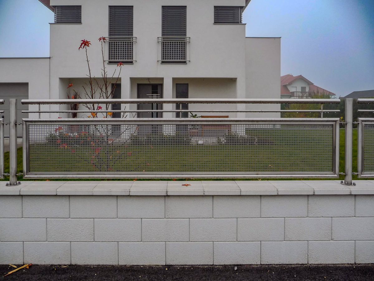 Detailaufnahme eines modernen Edelstahl-Zaunelements mit Lochblech-Füllung und Betonsockel, vor einem weißen Einfamilienhaus bei nebligem Wetter
