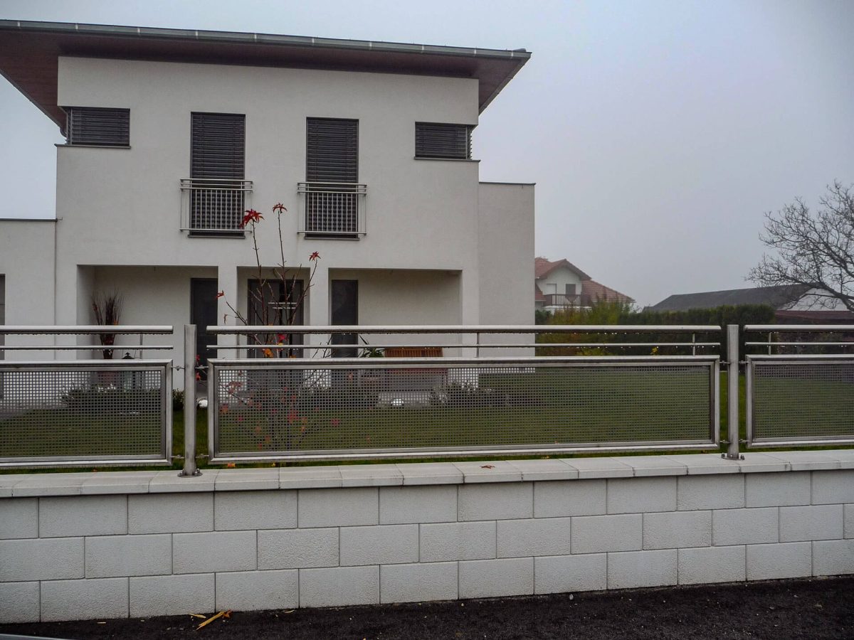 Moderner Gartenzaun aus Edelstahl mit Lochblech-Füllung und Betonmauer, vor einem weißen, zweigeschossigen Wohnhaus an einem nebligen Tag
