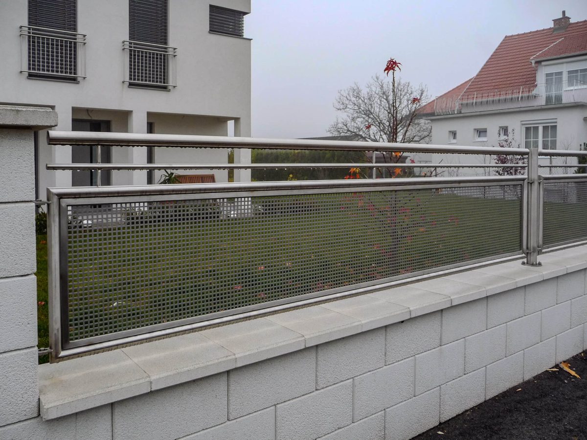 Horizontales Zaunelement aus Edelstahl mit gelochter Blechfüllung, montiert auf einer weißen Mauer vor einem modernen Wohnhaus