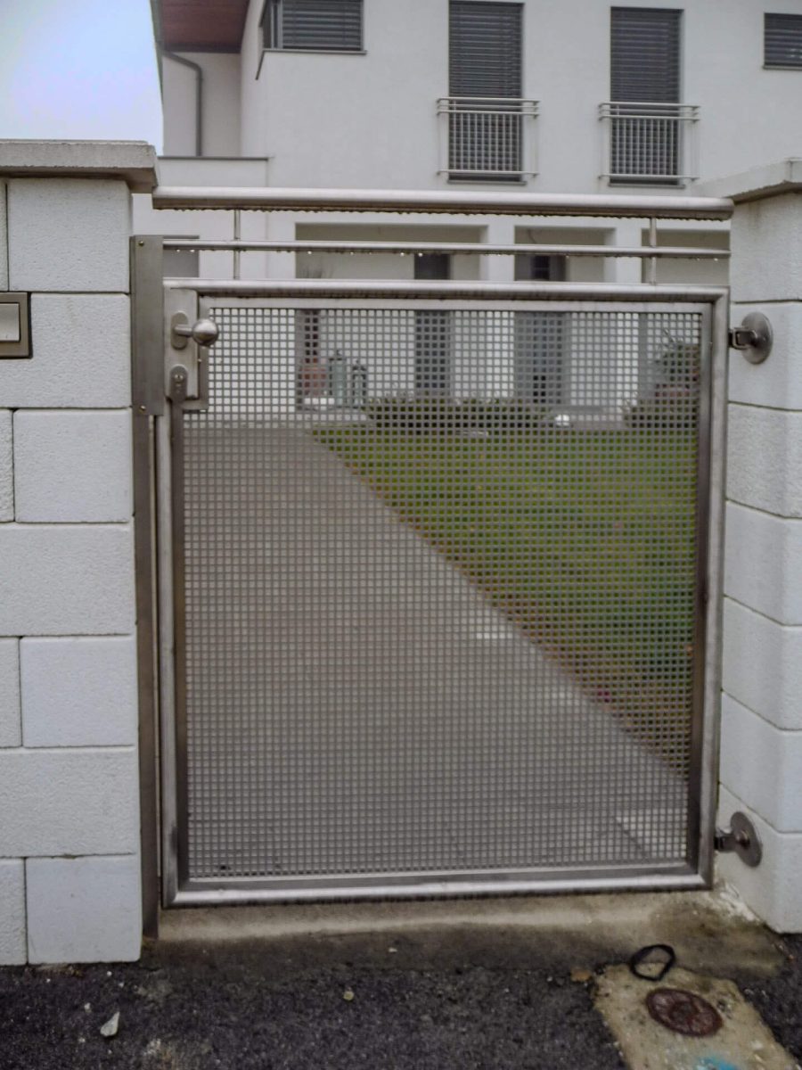 Modernes Eingangstor aus Edelstahl mit gelochter Blechfüllung und seitlich montierten Scharnieren, eingebaut in eine weiße Steinmauer vor einem Wohnhaus