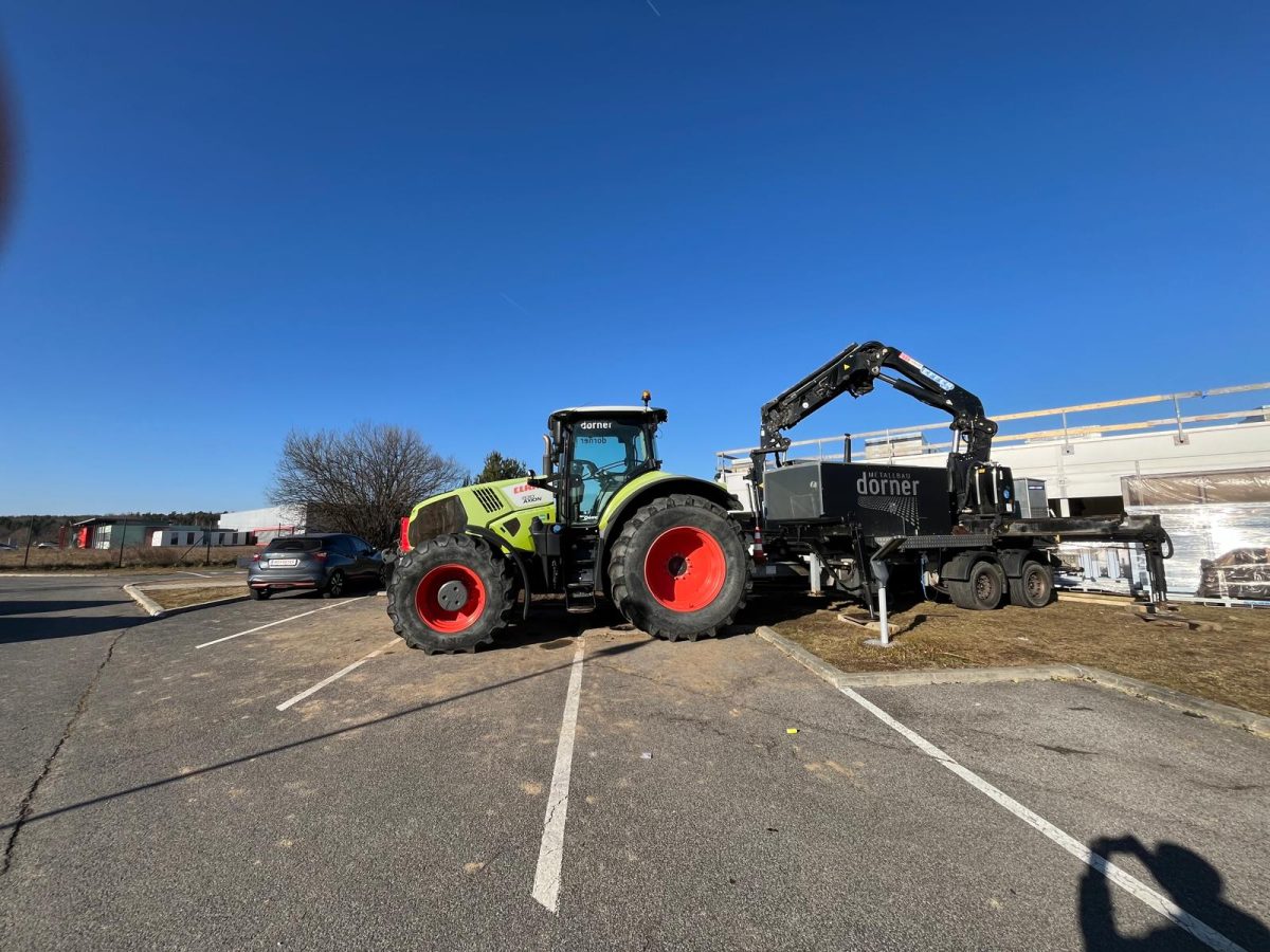 Grüner Traktor mit Anhänger und aufgebautem Kran der Firma Metallbau Dorner vor einem Rohbau bei strahlend blauem Himmel