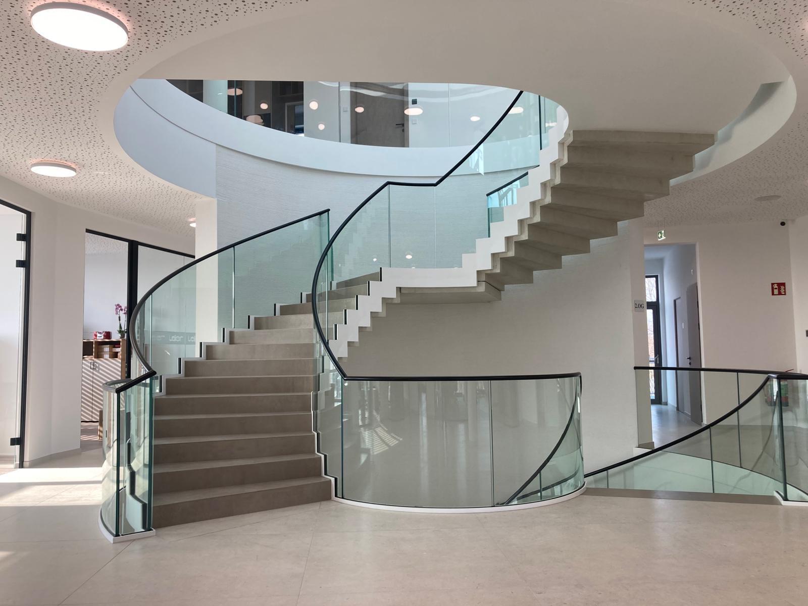Elegante geschwungene Treppe mit Glasbrüstung und dunklem Handlauf in lichtdurchflutetem Foyer