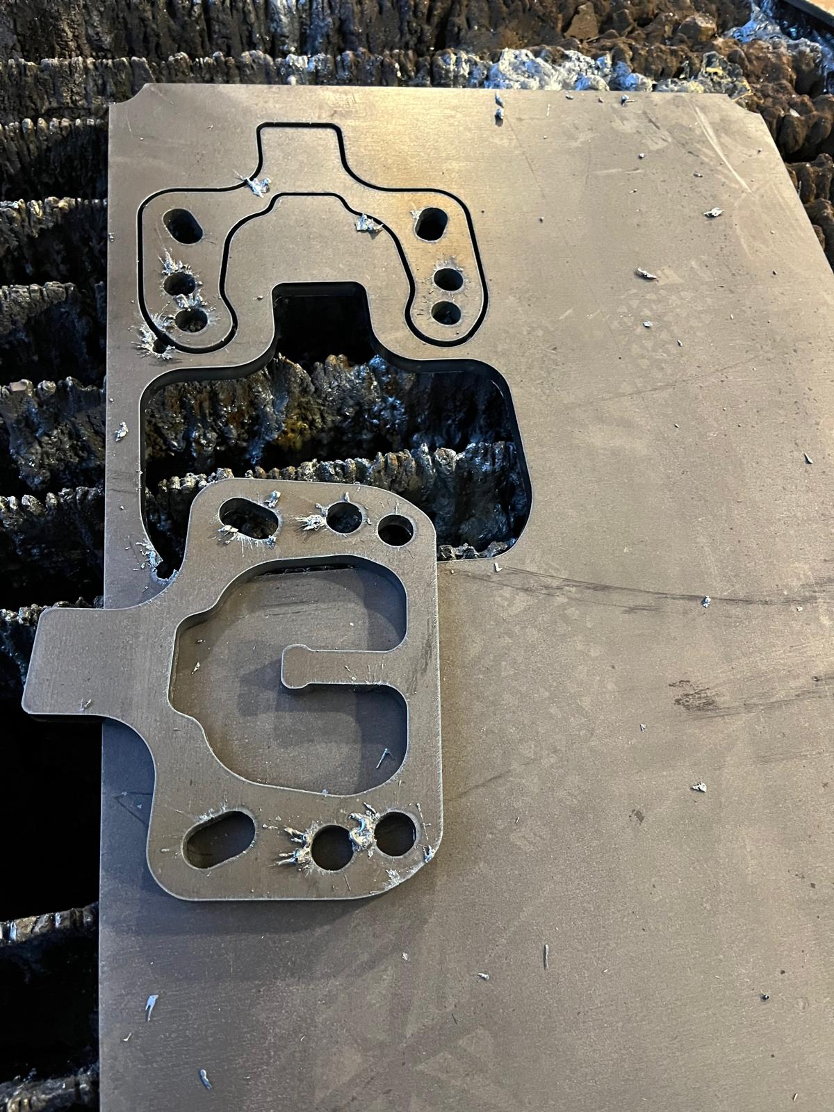 Zwei ausgebrannte Metallteile mit detaillierten Konturen und Bohrungen liegen auf einer Stahlplatte nach dem Plasmaschneiden