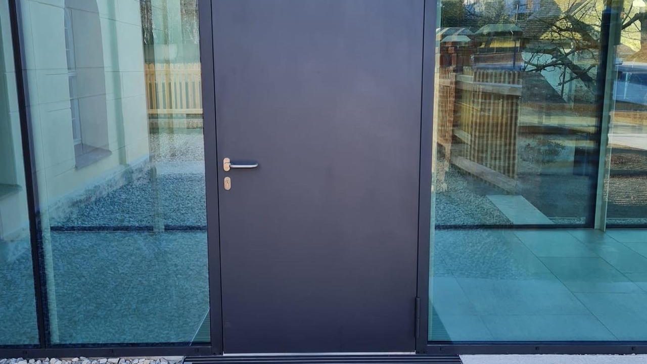 Dunkle Metalltür mit Griff, eingebaut zwischen zwei bodentiefen Glasflächen in einem modernen Eingangsbereich
