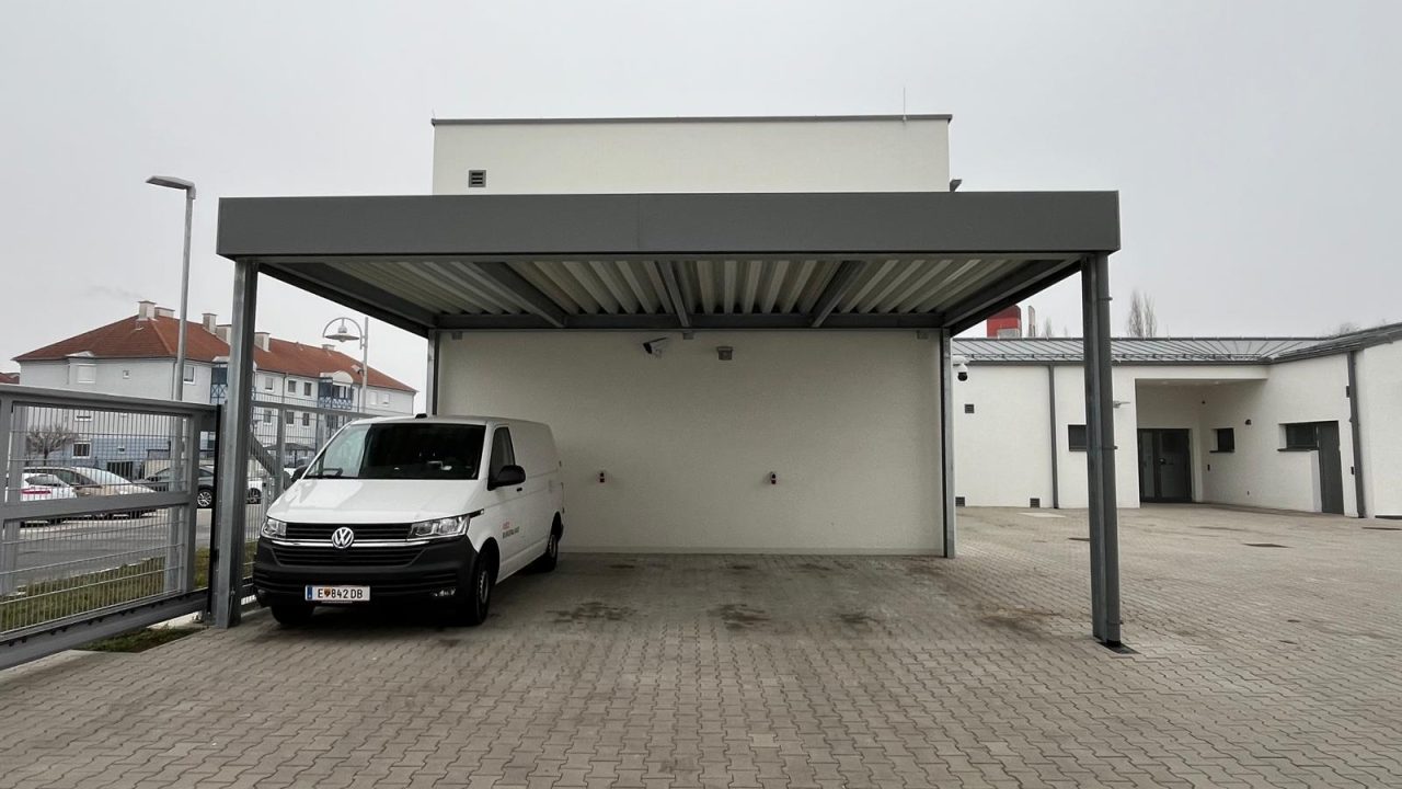 Großer Carport mit Stahlkonstruktion und Flachdach vor einem Firmengebäude, ein weißer Transporter ist darunter geparkt