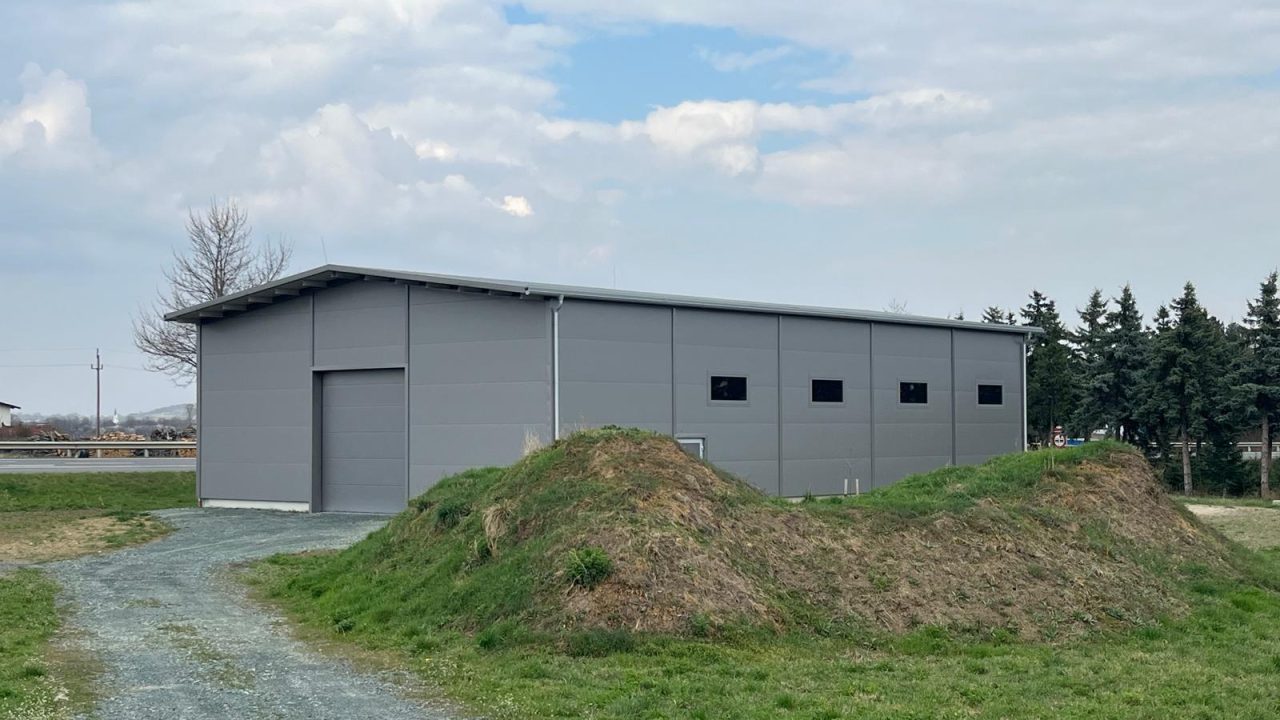 Freistehende Halle mit grauen Fassadenpaneelen, Satteldach und Zufahrtsweg auf einer Grünfläche