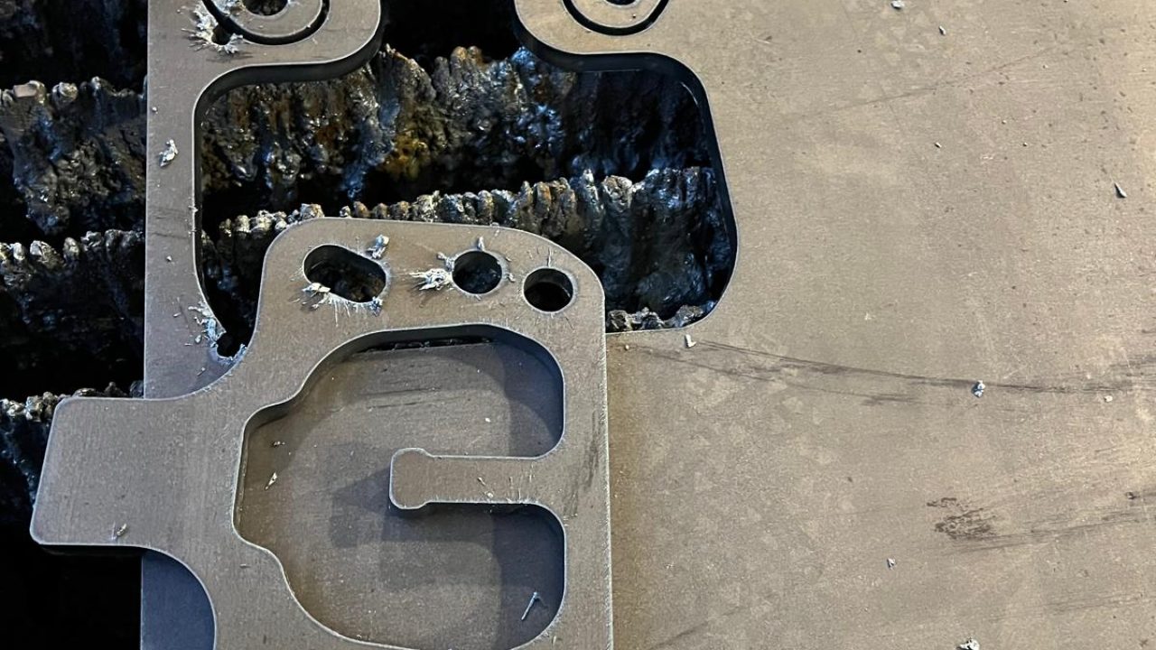 Zwei ausgebrannte Metallteile mit detaillierten Konturen und Bohrungen liegen auf einer Stahlplatte nach dem Plasmaschneiden