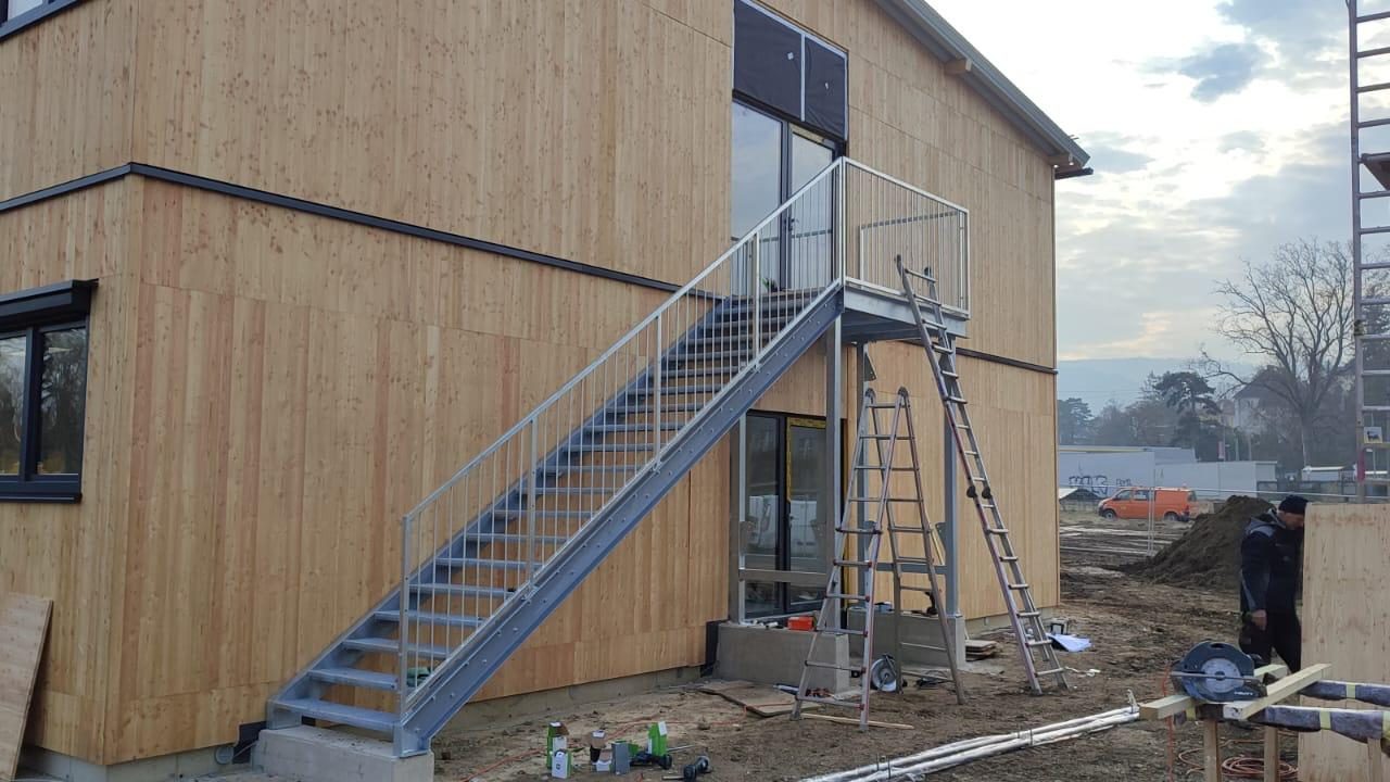 Geradläufige Stahltreppe mit Podest und Geländer an einem zweistöckigen Gebäude mit vertikaler Holzverkleidung