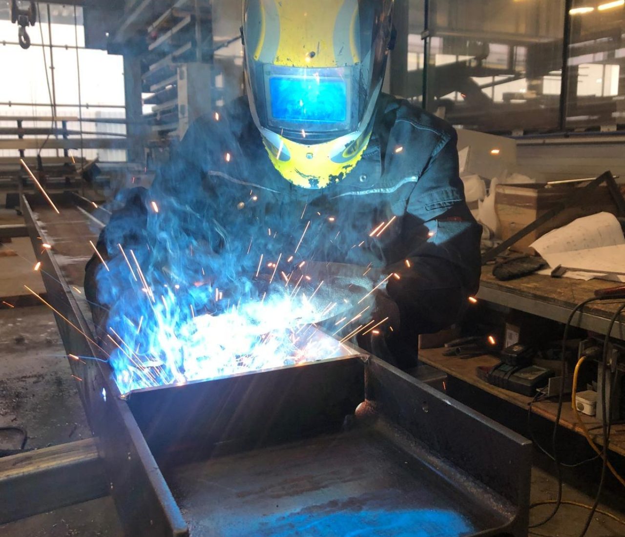 Schweißer mit gelbem Helm arbeitet mit hellen Lichtbögen an einem Stahlträger in einer Produktionshalle
