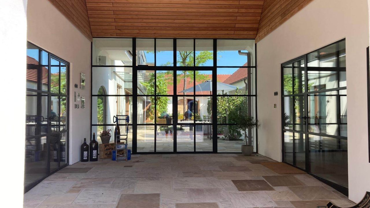 Überdachter Innenhof mit schwarzem Stahl-Glas-System, Natursteinplattenboden und Holzdecke. Blick auf offenen Innenhof mit Bepflanzung.