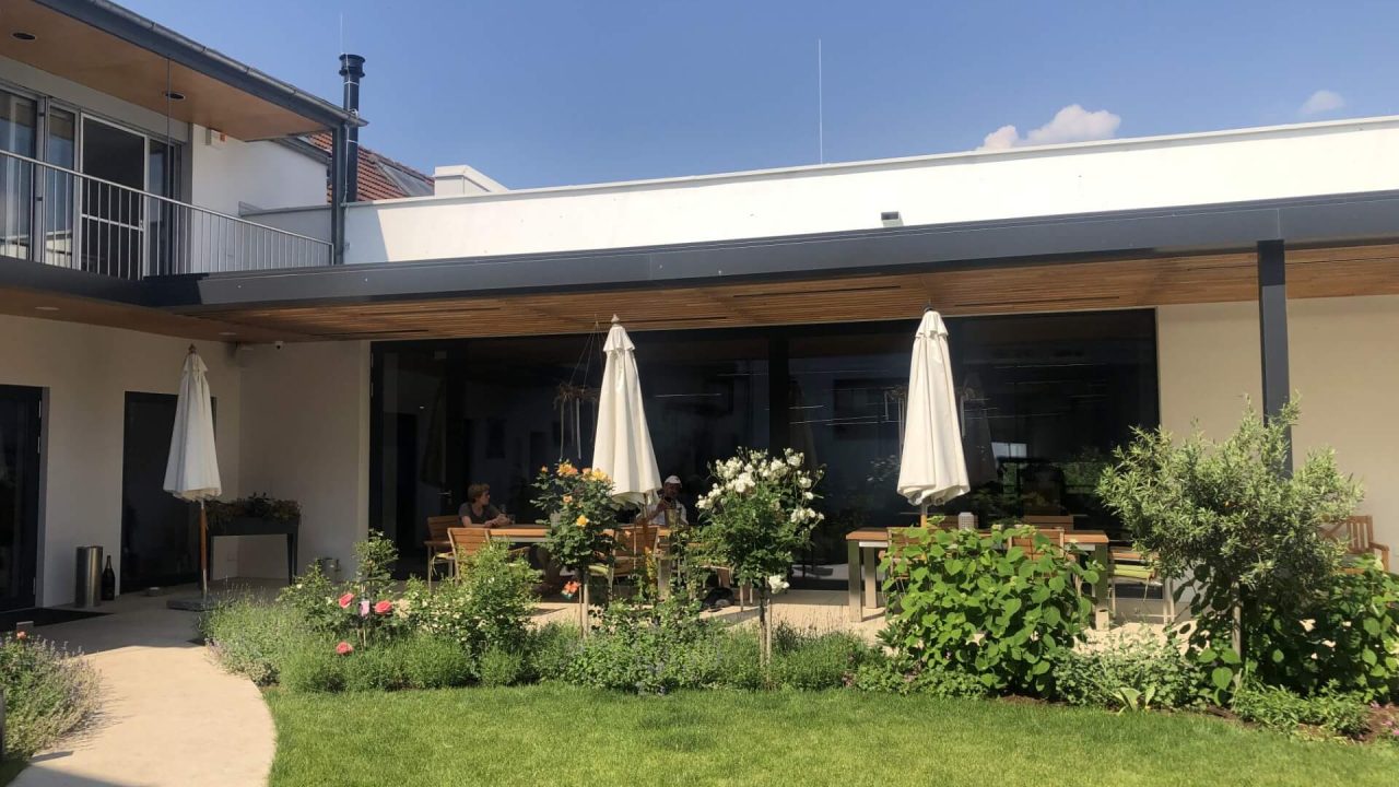 Grüner Innenhof mit gepflegtem Rasen, Blumenbeeten und überdachter Terrasse mit Sitzgruppen und Sonnenschirmen – ideal für entspannte Zusammenkünfte.