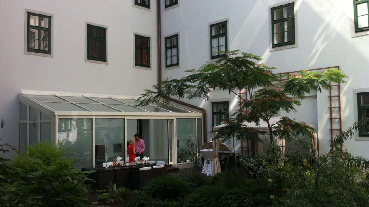 Moderner Wintergarten mit Glasdach in einem begrünten Innenhof, ideal für geschützten Aufenthalt im Freien bei jeder Witterung.